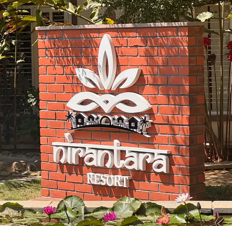 Brick entrance sign at Nirantara Resort & Spa with the resort's white logo, surrounded by greenery and water lilies